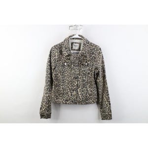 May include: A long-sleeve denim jacket featuring a tan and black leopard print pattern. The jacket includes a pointed collar, a front button-down closure, two chest pockets with buttoned flaps, and buttoned cuffs. A white brand label is visible inside.