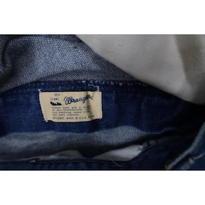 Op de afbeelding: Close-up van een donkerblauw denim kledingstuk met een zichtbaar label. Het label luidt "Wrangler" en bevat wasvoorschriften. Het kledingstuk is gemaakt in de VS.