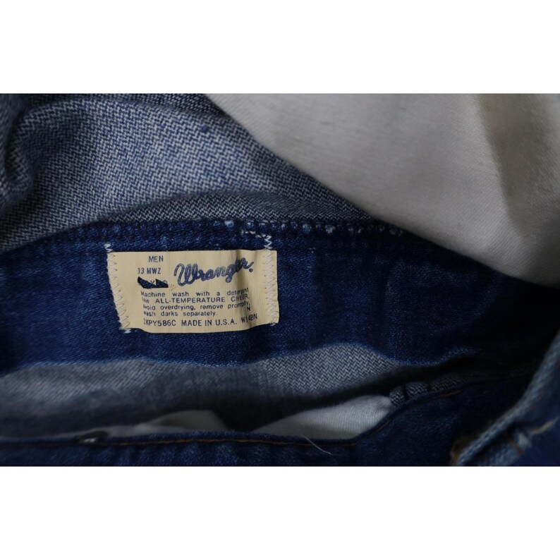 Op de afbeelding: Close-up van een donkerblauw denim kledingstuk met een zichtbaar label. Het label luidt "Wrangler" en bevat wasvoorschriften. Het kledingstuk is gemaakt in de VS.