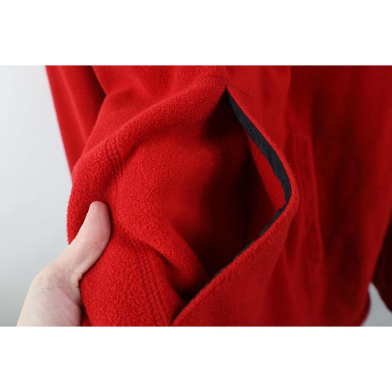 May include: Close-up of a red fleece jacket with a black-lined pocket. The soft, textured fabric is a vibrant red, and the pocket is visible on the left side. The jacket appears to be warm and comfortable.