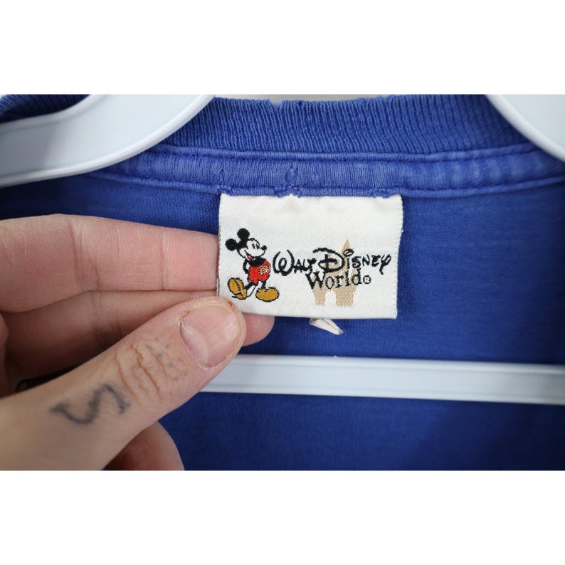 May include: A close-up of a blue t-shirt with a Walt Disney World tag. The tag features a cartoon Mickey Mouse and the words "Walt Disney World" in a stylized font. The shirt is hanging on a white hanger.