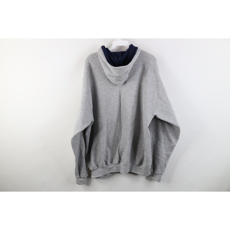 May include: A heather gray long-sleeve pullover hoodie featuring a contrasting dark navy blue interior lining within the hood. The garment is constructed with a standard drawstring-free hood, ribbed cuffs at the sleeves, and a ribbed hemline.