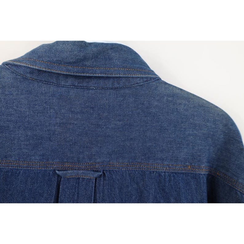 May include: Close-up of the back of a blue denim shirt. The collar and yoke are visible, with brown stitching. The fabric is a medium wash denim, and the shirt appears to be new or in excellent condition.