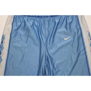 May include: Light blue athletic shorts featuring a white Nike swoosh logo on the right leg. The sides of the shorts have white panels decorated with a blue argyle diamond pattern outlined in black, separated by a thin dark blue vertical stripe.