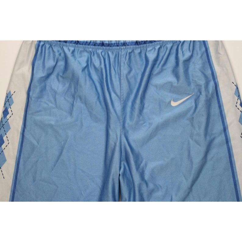 May include: Light blue athletic shorts featuring a white Nike swoosh logo on the right leg. The sides of the shorts have white panels decorated with a blue argyle diamond pattern outlined in black, separated by a thin dark blue vertical stripe.