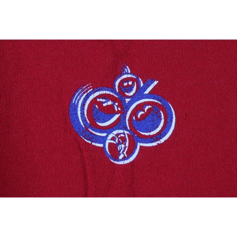 May include: A maroon-colored t-shirt featuring a distressed graphic design. The graphic is a series of connected, abstract shapes in blue and white. The design has a vintage, worn appearance.