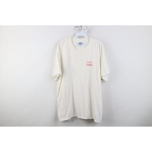 May include: Off-white short-sleeved t-shirt hanging on a white hanger. The shirt has the words "NOVEL" and "COMPANY" printed in red on the upper left chest area.