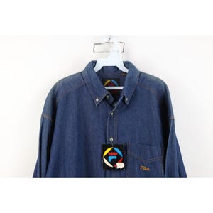 May include: A blue denim button-down shirt with a pocket on the left chest. The shirt has a collar and long sleeves. A tag with the Fila logo is attached. The Fila logo is also embroidered on the pocket.