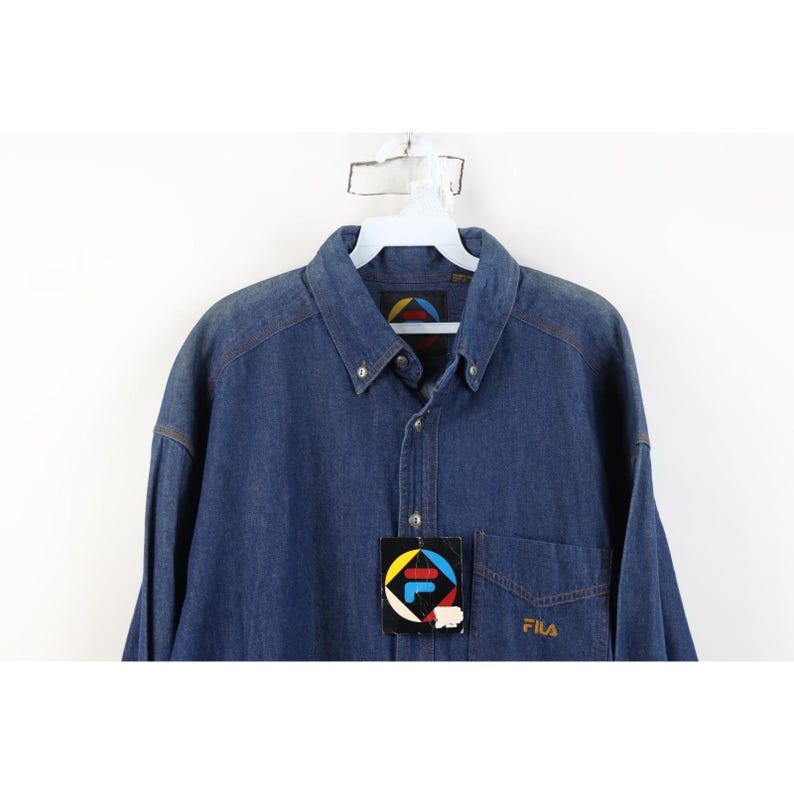 May include: A blue denim button-down shirt with a pocket on the left chest. The shirt has a collar and long sleeves. A tag with the Fila logo is attached. The Fila logo is also embroidered on the pocket.