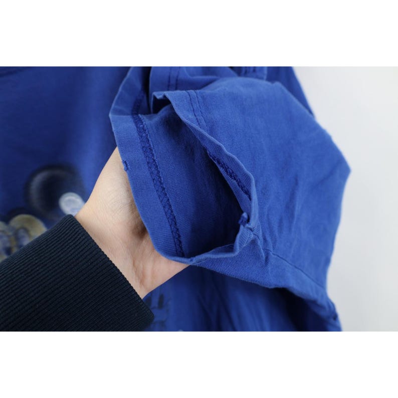 May include: A close-up of a royal blue t-shirt sleeve. The sleeve is held up, revealing the hem and stitching. The fabric appears to be a soft cotton material. The shirt has a faded design on the front.