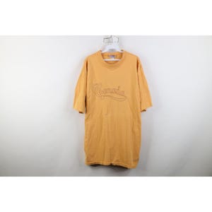 May include: Mustard yellow t-shirt with the word "Canada" embroidered across the chest in a cursive font. The shirt has short sleeves and a crew neck. The garment is hanging on a white hanger against a white background.