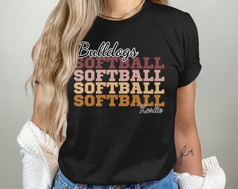 May include: Black t-shirt with the text "Bulldogs Softball" repeated four times in a gradient of brown, pink, and orange. The text "Karlie" is below the repeated text.