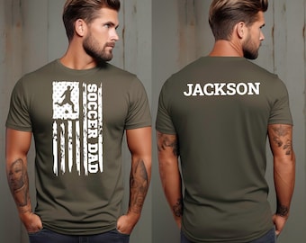 May include: A green t-shirt with a white distressed American flag design and the text "Soccer Dad". The back of the shirt has the text "JACKSON" in white.