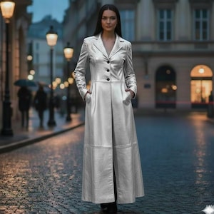 Könnte beinhalten: Ein langer, silberfarbener Mantel mit klassischem Reverskragen und schwarzen Knöpfen. Der Mantel hat Taschen und eine taillierte Passform, geeignet für Oberbekleidung. Das Model steht auf einer Kopfsteinpflasterstraße.