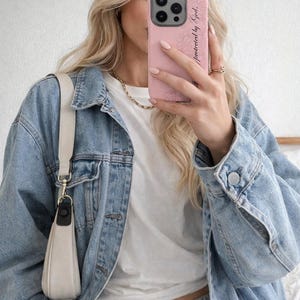 Könnte beinhalten: Hellblaue Jeansjacke, weißes T-Shirt und graue Jogginghose. Eine rosa Handyhülle mit dem Text "Protected by God" wird hochgehalten. Eine weiße Handtasche mit schwarzem Riemen hängt an der Schulter.
