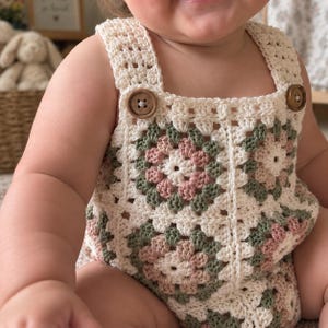 May include: A crochet romper featuring a granny square pattern in cream, sage green, and dusty rose. The garment has wide shoulder straps secured with two round brown wooden buttons at the front, showcasing a textured, handmade yarn construction.