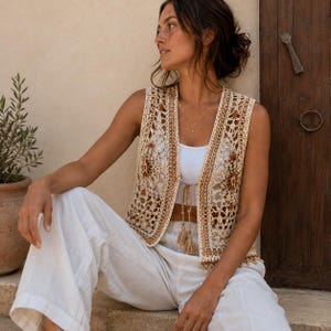 May include: A sleeveless crochet waistcoat featuring an intricate openwork pattern in cream and brown tones. The waistcoat has a V-neckline, a front tie closure with tassels, and a straight hem. It is worn over a plain white vest and white trousers.
