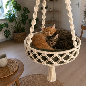 May include: A cream-colored macrame hanging cat bed featuring a circular frame with a diamond-patterned weave, suspended by three twisted ropes. Inside the bed, an orange tabby cat and a brown tabby cat are curled up together on a dark olive cushion.