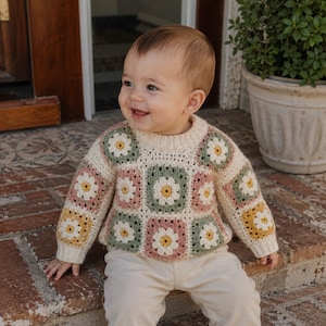 May include: A long-sleeve jumper featuring a crochet granny square pattern with daisy motifs in shades of cream, sage green, dusty pink, and mustard yellow. The jumper has a ribbed crew neckline and ribbed cuffs, constructed from yarn.