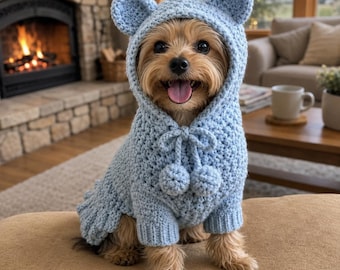 May include: A small dog with tan and brown fur wearing a light blue crocheted hooded sweater. The sweater features two small rounded ears on top of the hood, a textured knit pattern, and two dangling pom-poms attached to a drawstring at the neck.