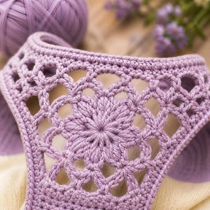 Könnte beinhalten: Nahaufnahme eines lila Häkelstücks mit einem floralen Lochmuster. Die Häkelarbeit ist detailliert, mit einem zentralen Blumenmotiv und einem offenen, spitzenartigen Muster. Im Hintergrund ist ein Knäuel lila Garn zu sehen, was auf ein handgemachtes Bastelprojekt hindeutet.