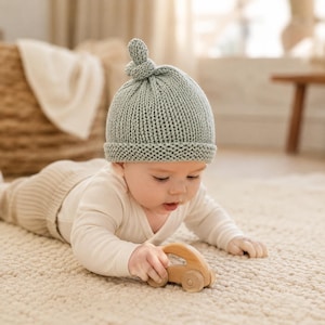 Può includere: Un cappello da neonato lavorato a maglia verde chiaro con un nodo sulla parte superiore. Il bambino indossa un body bianco a maniche lunghe e gioca con un'auto giocattolo di legno. Il bambino è sdraiato su un tappeto color crema.