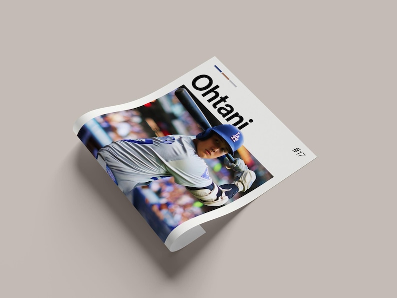 May include: A printed poster featuring a baseball player in a Dodgers uniform, mid-swing with a bat. The name "Ohtani" is printed in large black letters. The player is wearing a blue helmet with the Dodgers logo. The poster is on a light gray surface.