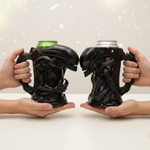 May include: Two black Alien-themed mugs, each with a detailed sculpted design of the creature's head and body. The mugs have large handles and are holding beverage cans. The mugs are held up by hands.