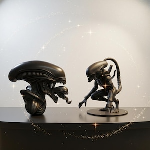 May include: Two black Alien figures are displayed on a dark surface. One is a bust with a detailed head and open mouth. The other is a full figure in a crouching pose, with a long tail and sharp claws. The background is a light color with sparkling effects.