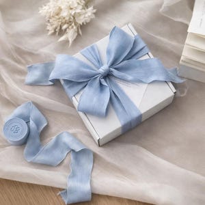 May include: A white gift box tied with a light blue ribbon and bow. A roll of matching ribbon with a circular emblem is unraveled on a soft, neutral-toned surface. Dried flowers and a stack of papers are in the background.