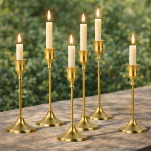 May include: Six gold-colored candle holders with lit, ivory-colored candles. The candle holders have a classic design with a wide base and a slender stem. The candles are positioned at varying heights, creating a visually appealing arrangement. The background is a blurred green.