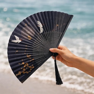 May include: A black folding hand fan with a crane and floral design. The fan is held open, revealing the intricate artwork. A black tassel hangs from the base of the fan. The background is a blurred view of the ocean.
