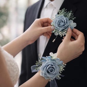 May include: A blue rose boutonniere and wrist corsage set. The boutonniere is pinned to a black suit jacket. The wrist corsage features a large blue rose with white accents and a blue ribbon. The set is ideal for weddings and formal events.