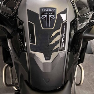 Puede incluir: Depósito de combustible de motocicleta negro y plateado. Presenta los textos "TIGER 1200" y "GT EXPLORER". Un panel negro texturizado tiene un diseño de garra y la frase "FOR THE RIDE".