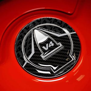May include: A close-up of a black and silver motorcycle fuel cap with the text "V4 STREETFIGHTER" on a red surface. The cap features a carbon fibre pattern and a silver V4 emblem.