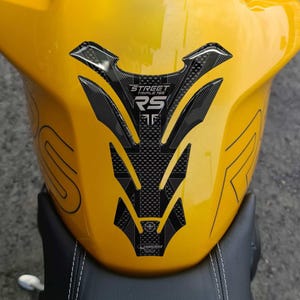 Puede incluir: Un depósito de gasolina amarillo de motocicleta con un protector de depósito negro con textura de fibra de carbono. El protector tiene el texto "STREET TRIPLE 765 RS" y un logotipo. El asiento es negro.