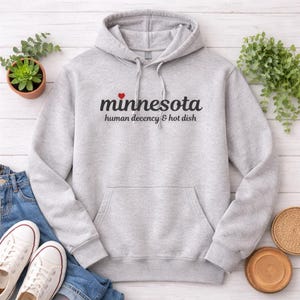 May include: A light gray hooded sweatshirt with the word "minnesota" in black script, a red heart, and the text "human decency & hot dish." The sweatshirt has a front pocket and a drawstring hood. White sneakers and blue jeans are visible.