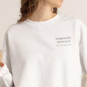 May include: A white crewneck sweatshirt featuring the black text "temporarily possessed" on the upper left chest, with the smaller text "please try again later." printed directly underneath the main phrase in a simple, clean font style.