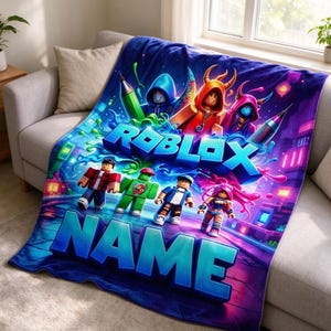 May include: A soft plush throw blanket featuring a vibrant Roblox graphic with multiple block-style characters, neon green paint splatters, and large blue 3D text that reads ROBLOX and NAME. The design includes a dark purple and blue background.