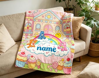 May include: A pink blanket featuring a whimsical design with cartoon bears, a rainbow, and a house-shaped structure. The blanket has a personalized "name" text, cupcakes, and lollipops. The blanket is draped over a beige sofa.