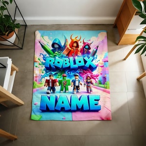 May include: A rectangular fleece blanket featuring a vibrant Roblox-themed graphic. The design displays various blocky character figures, colorful paint splashes, and the bold blue text ROBLOX and NAME printed in a large, stylized font.