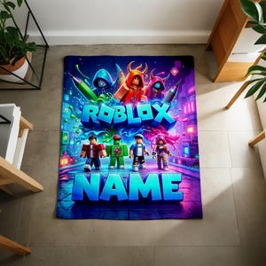 May include: A rectangular blanket featuring a vibrant Roblox-themed graphic with four blocky character figures, colorful paint splatters, and the text ROBLOX and NAME in large blue letters against a dark, neon-lit digital background.