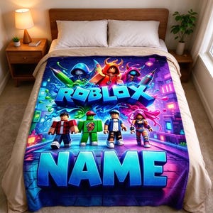 May include: A rectangular fleece blanket featuring a vibrant Roblox graphic with multiple block-style characters, colorful paint splatters, and the text ROBLOX and NAME in large blue letters, displayed spread out over a bed with white pillows.