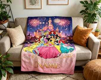 May include: A pink and blue blanket featuring a castle scene with fireworks and several cartoon princesses. The blanket has the word "NAME" in gold script at the bottom. The blanket is draped over a beige sofa.