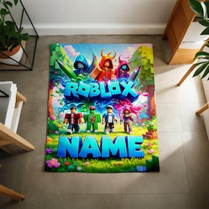 May include: A rectangular blanket featuring a vibrant Roblox-themed graphic with blocky characters, colorful splashes, and the text ROBLOX NAME in large blue letters. The design includes a grassy landscape with trees and flowers.
