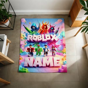 May include: A rectangular area rug featuring a vibrant Roblox graphic with the text ROBLOX and NAME in large 3D letters. The design includes four blocky character figures, colorful paint splashes, and building blocks in shades of blue, pink, and green.