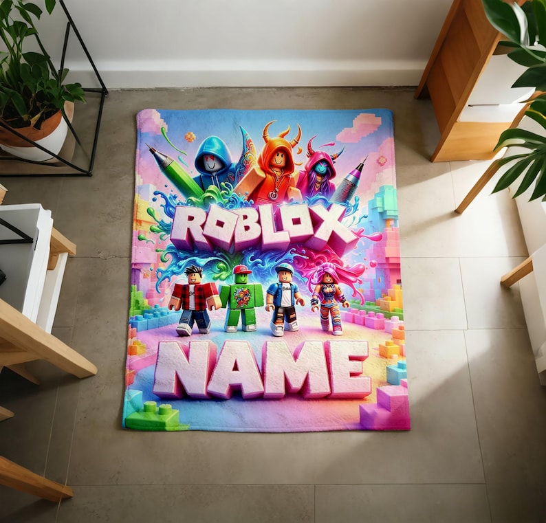 May include: A rectangular area rug featuring a vibrant Roblox graphic with the text ROBLOX and NAME in large 3D letters. The design includes four blocky character figures, colorful paint splashes, and building blocks in shades of blue, pink, and green.