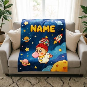 Pode incluir: Uma manta de lã azul macia com um tema espacial, incluindo estrelas amarelas, planetas, um foguetão e uma grande lua amarela. No centro, uma personagem de desenho animado com uma cabeça de máquina de pastilhas sopra uma bolha cor-de-rosa.