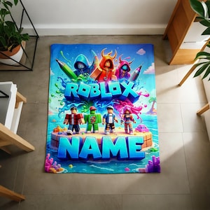 May include: A rectangular blanket featuring a vibrant Roblox-themed graphic with the text ROBLOX and NAME in bold blue letters. The design includes four blocky character figures standing on a sandy island surrounded by blue water and colorful splashes.