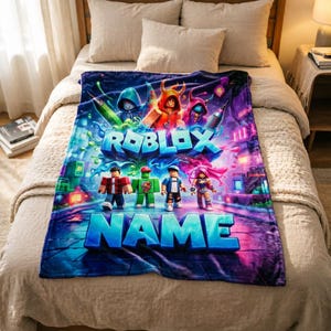 May include: A vibrant blanket featuring the Roblox logo and characters, set against a colourful cityscape. The word "NAME" is displayed at the bottom. The blanket is shown on a bed with neutral-toned bedding.